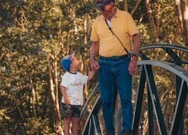 A grandfather and grandson enjoying a sunny day walk on a rustic bridge surrounded by trees.